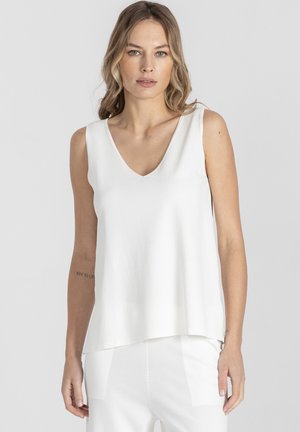 Kobieta z falującymi blond włosami, ubrana w białą bezrękawnikową bluzkę z dekoltem w serek i białe spodnie, stojąca na tle jasnoszarej ściany.