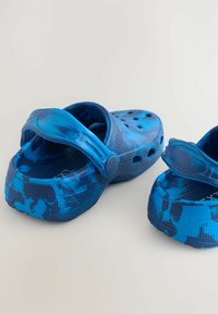 Zila plastmasas klumpes ar kamuflāžas rakstu, atvērtu dizainu ar ventilācijas caurumiem, regulējamu siksnu un tekstūrētiem zoliņām labākai saķerei.