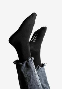 Schwarze Baumwollsocken mit glatter Textur, versehen mit einem in Weiß bedruckten Etikett. Getragen mit ausgefransten, hochgekrempelten Jeans für einen lässigen Stil.
