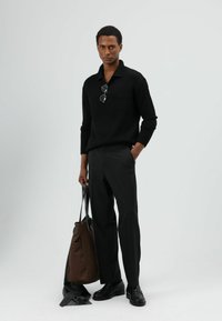 Zwarte gebreide trui, donkere op maat gemaakte broek, zwarte schoenen, met een bruine draagtas in de hand. Bril ligt op de kraag van de trui. Eenvoudig en minimalistisch ontwerp.