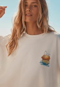 Sweatshirt blanc avec un design brodé d'un citron et de Snoopy. Le tissu semble doux, avec une coupe décontractée et un col rond.