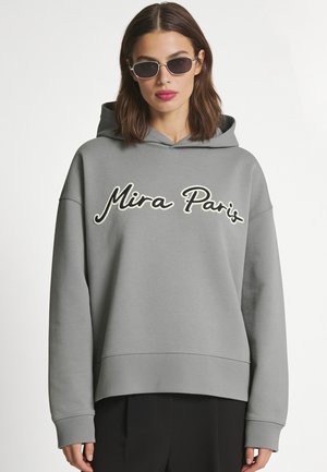 Vrouw die een grijze "Mira Paris" capuchontrui en donkere zonnebril draagt, staand tegen een effen witte achtergrond.