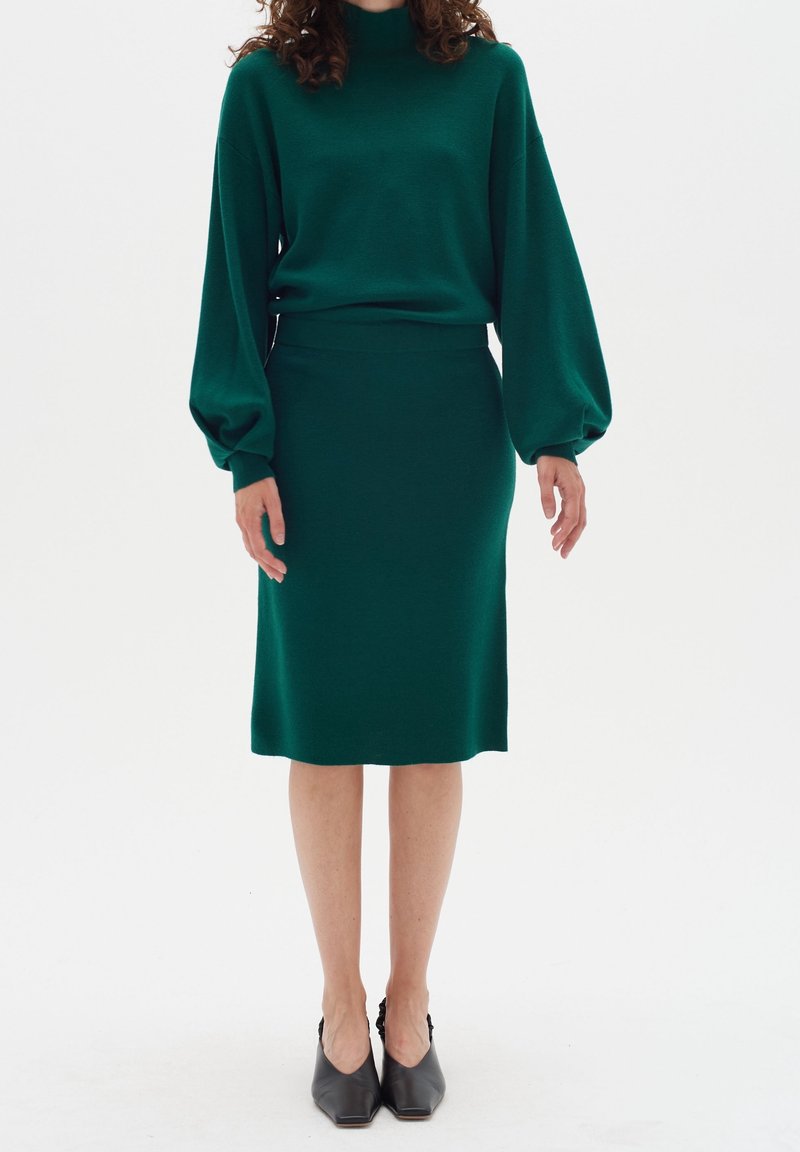 Femme portant un pull à manches longues vert foncé avec des manches bouffantes et une jupe assortie jusqu'aux genoux, associée à des chaussures noires à bout pointu.