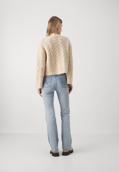 Pulover bej din tricot cu un model texturat în zigzag, asortat cu blugi deschiși la culoare și evazați, împreună cu încălțăminte închisă la culoare, văzut din spate.