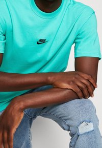 Turkos bomull t-shirt med svart Nike-logga på bröstet, korta ärmar och rund hals. Modellen bär ljusblå slitna jeans.