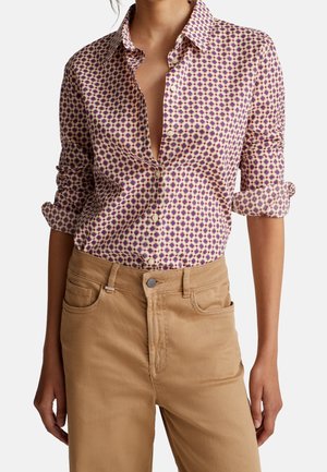 Vrouw draagt een zijden blouse met lange mouwen en patroon, mouwen opgerold en beige broek met hoge taille, staand met ontspannen handen.