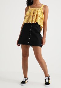 Gul blommig ärmlös topp med lager av volanger; svart denimkjol med knappar fram och fransig nederkant; svarta och vita mönstrade sneakers.