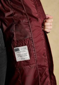 Burgundi värvi pehme voodriga jakk, millel on sisemine tasku ning silt hooldusjuhiste ja brändi üksikasjadega.