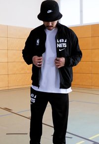 Giacca nera Nike con logo NBA Nets, maglietta bianca sotto, e pantaloni della tuta neri con logo. Indossata con un cappello a secchiello nero. Ambiente interno.