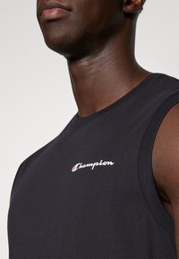 Camiseta de algodón sin mangas de color negra con un logo bordado en blanco de "Champion" en el área del pecho izquierdo. Textura suave y diseño casual.