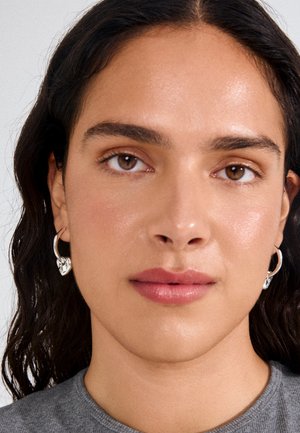 Ruskeasilmäinen ja tummatukkainen nainen käyttää hopeisia renkkikorvakoruja ja harmaata toppia, katsoo rauhallisesti kameraan neutraalia taustaa vasten.