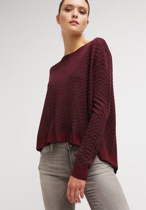 Maglione a maglia bordeaux con motivo a zigzag, polsini e orlo a coste, e una vestibilità rilassata, abbinato a jeans grigi chiari.