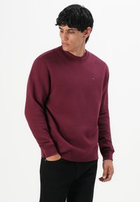 Joven vestido con un suéter burdeos de manga larga y pantalones negros, mirando hacia abajo a su izquierda con una mano en el bolsillo.