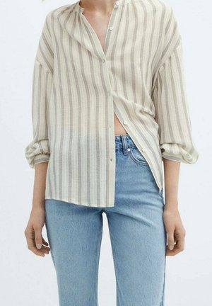Person iført en løs beige og hvidstribet skjorte med knapper, med ærmerne rullet op, og blå højttaljede jeans, stående mod en ensfarvet baggrund.