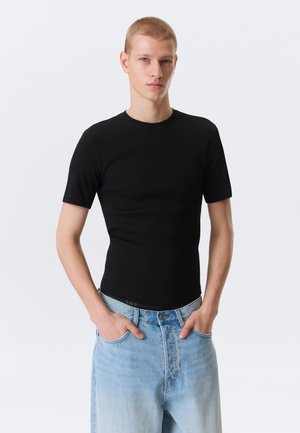 Giovane con capelli biondi corti, indossa una maglietta nera aderente a maniche corte e jeans di denim azzurro chiaro, con le mani nelle tasche.