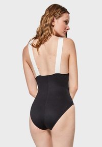 Bañador negro de una pieza con amplias tiras blancas, un diseño de espalda baja y textura suave. Presenta una silueta ajustada y costuras simples.
