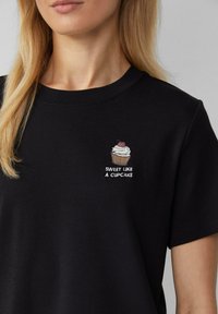 T-shirt en coton noir avec un graphique de cupcake avec de la crème et une cerise, et le texte "DOUX COMME UN CUPCAKE" en blanc en dessous. Manches courtes.