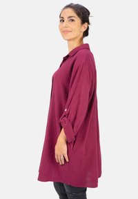 Maroon button-up shirt with a textured weave, long sleeves rolled at the cuffs, and a relaxed fit. Worn over black pants.