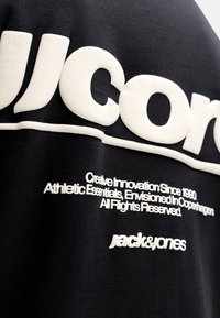 Zwarte trui met grote witte blokletters en kleinere witte tekst. De stof is katoen met een zachte textuur. Geaccentueerd met het merklogo.