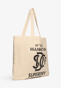 Beżowa torba na ramię z płótna z dwoma uchwytami, z czarnym nadrukiem: "34TH ST, MANHATTAN, SUPERDRY." Prostokątny kształt, wytrzymały materiał.