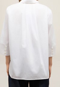 Chemise blanche avec un empiècement au dos, manches longues et poignets à boutons. Présente une texture lisse et une coupe décontractée.