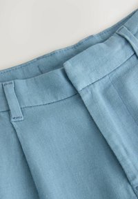 Pantalon en lin bleu clair avec une ceinture plate, des détails de couture visibles et une texture lisse, mettant en avant un design décontracté.