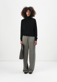 Un pull noir à col roulé, un pantalon gris à jambes larges, tenant un sac gris, avec une texture lisse et une coupe décontractée, sur fond blanc.