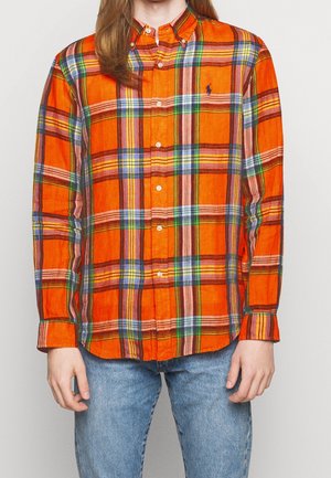 Mężczyzna w jasnopomarańczowej koszuli na guziki w kratę z niebieskimi, zielonymi i żółtymi paskami, w parze z jasnoniebieskimi jeansami.