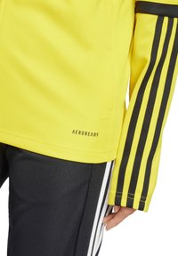 Gelbe Sportjacke mit strukturierter Oberfläche und schwarzen Streifen an den Ärmeln. "AEROREADY"-Logo am Saum. Schwarze Hose mit weißen Streifen.