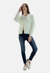 Weißer gestrickter Cardigan mit V-Ausschnitt und Bindeschnürung, getragen über einem grauen Top und kombiniert mit dunklen Jeans und weißen Sneakern.