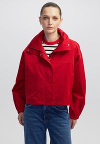 Vrouw met slicked-back haar, gekleed in gouden oorringen, een rode jas over een zwart-wit gestreepte blouse en blauwe jeans tegen een effen achtergrond.