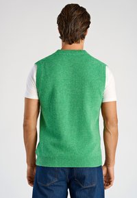 Grön väst i ullblandning med texturerad yta, ribbad fåll och ärmhål. Bärs över en vit t-shirt, vilket ger en avslappnad stil.