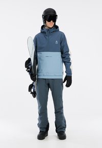 Snowboarder u plavoj i sivoj vodonepropusnoj jakni s kapuljačom i odgovarajućim hlačama. Crne rukavice i čizme, drži snowboard.