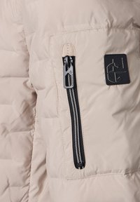 Beige gesteppte Jacke mit einer vertikalen schwarzen Reißverschlusstasche und einem schwarzen quadratischen Logopatch auf der Vorderseite.