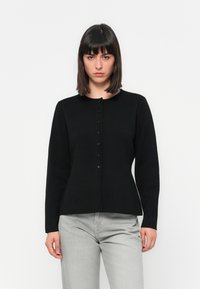 Kvinne med rett, mørkt hår, iført en tettsittende svart cardigan med knapper og lysegrå bukser, stående mot en enkel hvit bakgrunn.