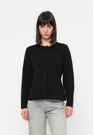 Femme avec des cheveux noirs raides portant un cardigan noir ajusté à boutons et un pantalon gris clair, se tenant contre un fond blanc uni.