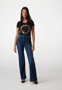 Sort tætsiddende t-shirt med grafisk print, kombineret med medium høj flared blå jeans, hvilket skaber et afslappet look. Modellen har sorte højhælede sko på.