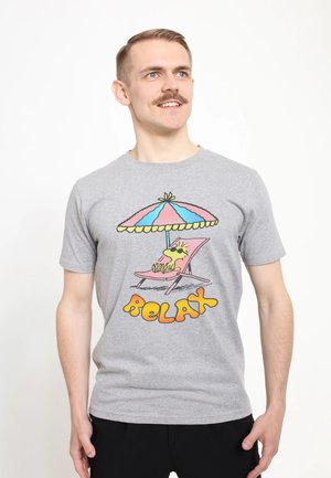 Mann mit Schnurrbart, der ein graues T-Shirt trägt und einen gezeichneten entspannenden Hund unter einem Sonnenschirm zeigt mit dem Schriftzug „Relax“ in Orange und Gelb.