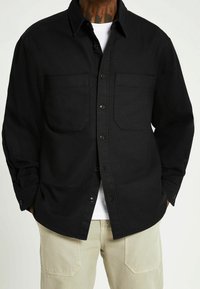 Czarna koszula z dwoma kieszeniami na piersi, zapinana na guziki, z długimi rękawami, wykonana z teksturowanej tkaniny. Nałożona na białą koszulkę.