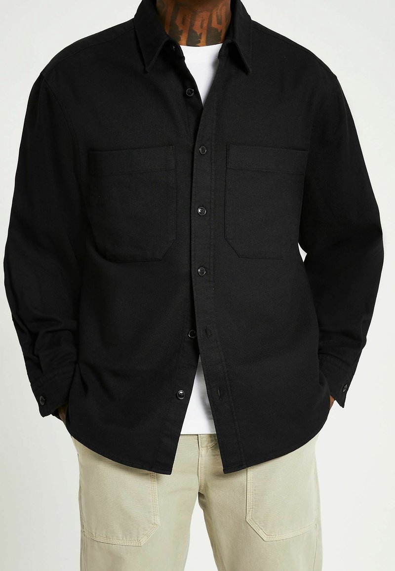 Czarna koszula z dwoma kieszeniami na piersi, zapinana na guziki, z długimi rękawami, wykonana z teksturowanej tkaniny. Nałożona na białą koszulkę.