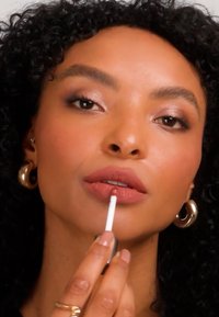 Kvinne med krøllete svart hår og gylne hoop-øredobber som påfører nude-farget leppeglans på leppene med en applikator.