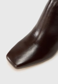 Primer plano de una bota de cuero marrón oscuro con una textura suave y punta cuadrada sobre un fondo blanco.