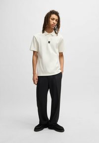 Weißes Polo-Shirt mit einer Vorderknopfleiste, schwarzem Logo-Patch auf der Brust, kombiniert mit lockeren schwarzen Hosen und schwarzfarbener Fußbekleidung.