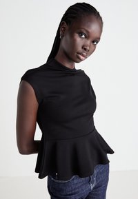 Young Black woman with braided hair wearing a sleeveless black peplum top and dark blue jeans against a plain white background.