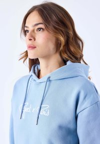 Sweat à capuche bleu clair en tissu doux. Comprend un texte brodé blanc "Project x Paris" et des œillets argentés pour les cordons.