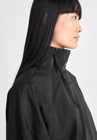 Schwarze Jacke mit hohem Kragen und glatter, wasserdichter Textur. Verfügt über einen Reißverschluss und ein minimalistisches Design mit subtilen Branding-Details.