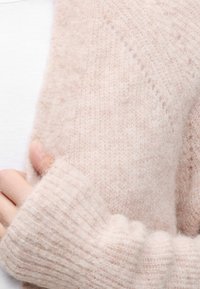 Personne portant un pull en maille doux et pelucheux de couleur rose pâle avec des poignets côtelés, tenant le tissu près de la poitrine par-dessus une chemise blanche.
