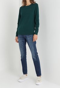 Vihreä neulepusero pyöreällä pääntiellä, yhdistetty sinisiin farkkuihin. Malli käyttää kermanvärisiä slip-on-tennareita. Yksinkertainen, rento asu.