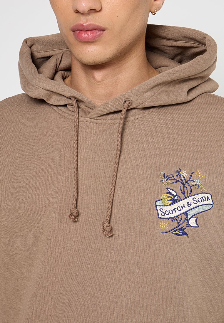 Bruine hoodie met een trekkoord, voorzien van een geborduurd bloemontwerp en de tekst "Scotch & Soda" op de voorkant.