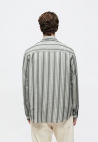 Hombre con cabello rizado castaño, vistiendo una camisa de manga larga a rayas verticales en gris y blanco y pantalones beiges, visto desde atrás.